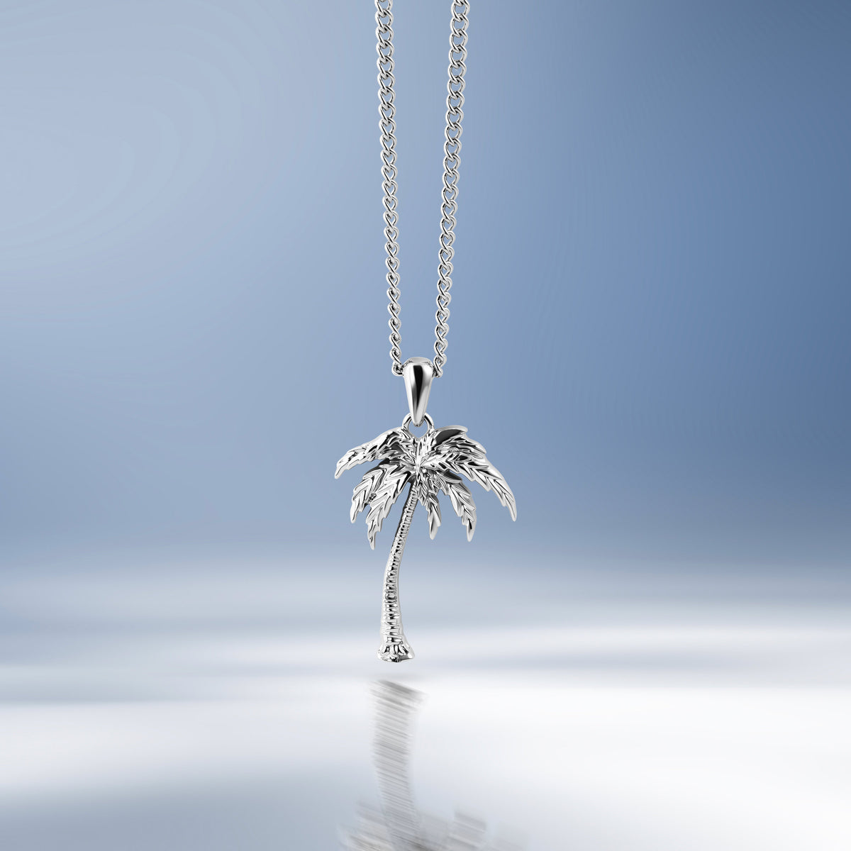 A silver palm tree pendant necklace hanging from a delicate silver chain against a soft blue gradient background with a reflective surface below.