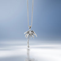 A silver palm tree pendant necklace hanging from a delicate silver chain against a soft blue gradient background with a reflective surface below.
