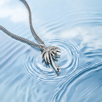 A silver-toned necklace with a detailed palm tree pendant, resting on rippling light blue water.