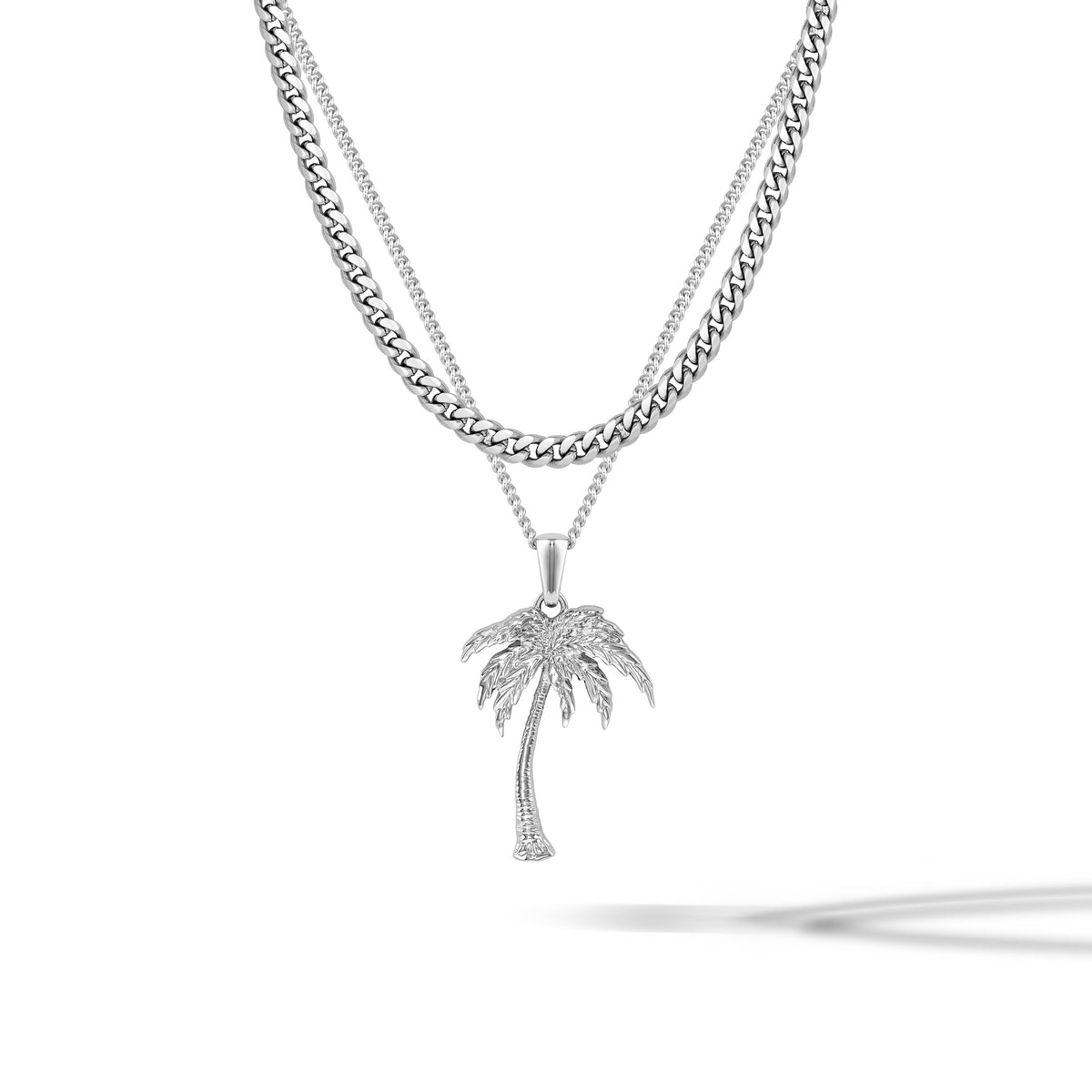 A silver-toned necklace featuring a double-strand chain design, with a curb chain on the top layer and a delicate chain on the bottom layer. Hanging from the bottom of the chain is a palm tree pendant adorned with small clear stones, set against a clean white background with a soft shadow.