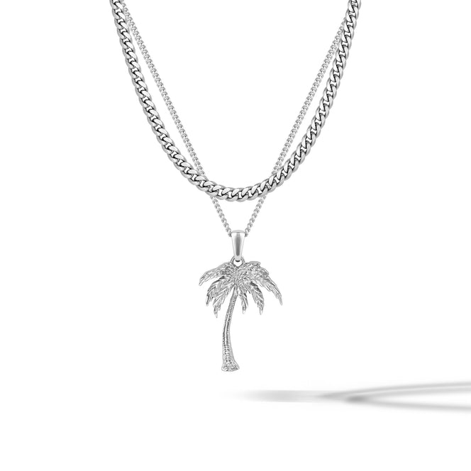A silver-toned necklace featuring a double-strand chain design, with a curb chain on the top layer and a delicate chain on the bottom layer. Hanging from the bottom of the chain is a palm tree pendant adorned with small clear stones, set against a clean white background with a soft shadow.