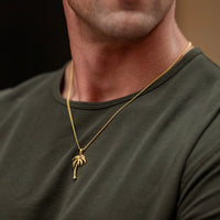 A close-up of a man wearing a gold chain necklace with a small palm tree pendant, paired with an olive green crewneck t-shirt.