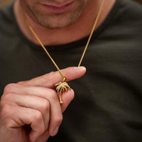 A close-up lifestyle shot of a man holding a small gold palm tree pendant on a gold chain necklace against his chest.