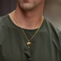 A close-up of a man wearing a gold chain necklace with a small palm tree pendant, paired with an olive green crewneck t-shirt.