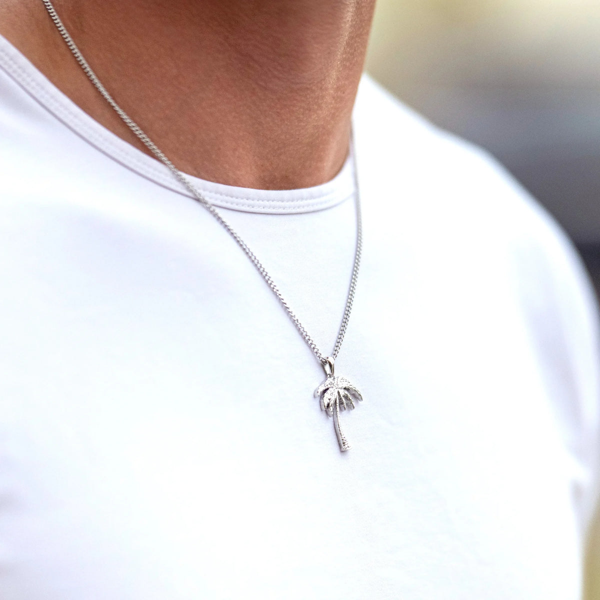 A close-up of a person wearing a silver chain necklace with a small palm tree pendant, styled with a white crew-neck t-shirt.