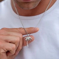 A close-up of a man wearing a silver chain necklace with a small palm tree pendant, held between his fingers against a white t-shirt.