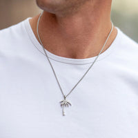 A close-up of a man wearing a silver chain necklace with a small palm tree pendant, paired with a white t-shirt.