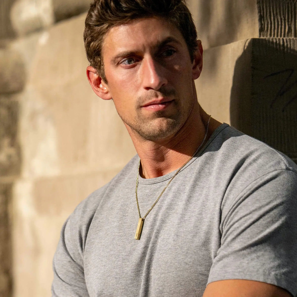 Man wearing a gray t-shirt with a necklace against a stone wall.