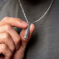 Hand holding a silver necklace with a rectangular pendant against a gray background