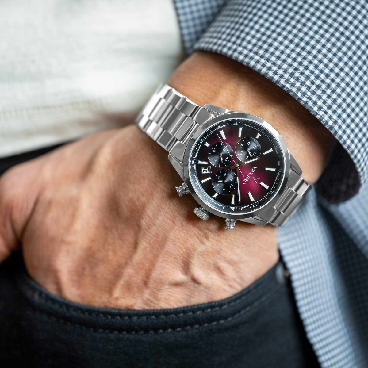 Silver wristwatch with a dark face on a person's wrist, wearing a blue checkered shirt.