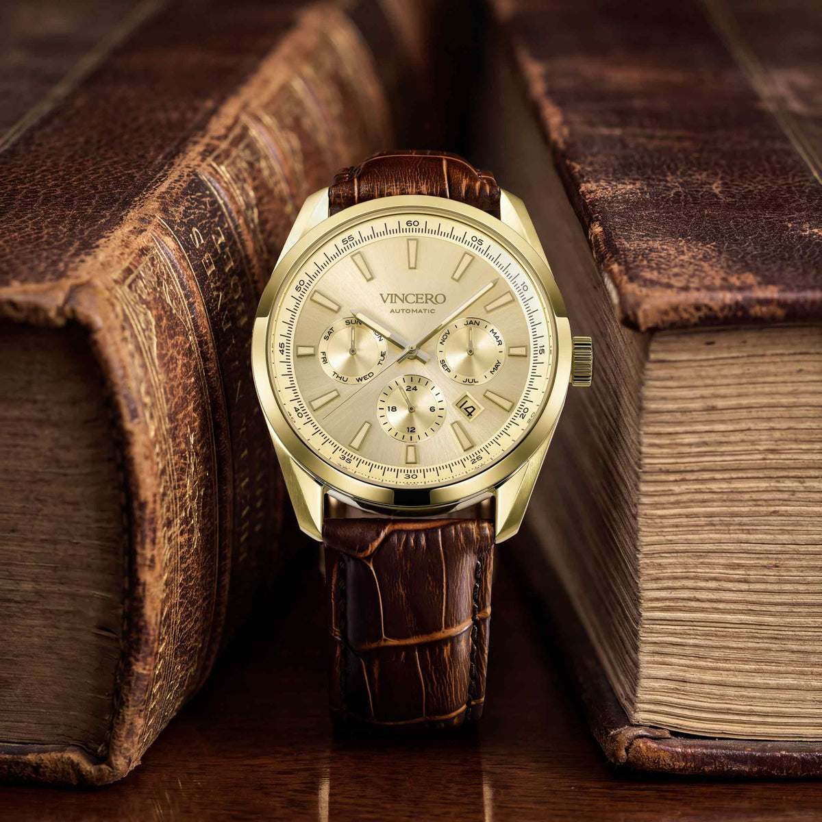 A VINCERO men's watch with a gold-tone case, beige dial, and brown leather strap, styled on a wooden surface between two vintage leather-bound books.