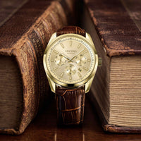 A VINCERO men's watch with a gold-tone case, beige dial, and brown leather strap, styled on a wooden surface between two vintage leather-bound books.