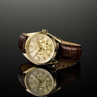 A Vincero wristwatch with a gold-toned case, champagne dial, and brown leather strap, displayed on a reflective black surface against a dark gray gradient background.