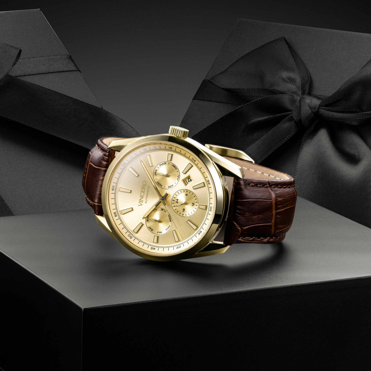 A luxury wristwatch with a gold-toned case, gold dial, and brown leather strap, displayed on a black pedestal in front of a black gift box with a bow.
