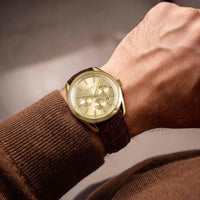 A close-up of a wrist wearing a gold-tone chronograph watch with a beige dial and a dark brown crocodile-embossed leather strap, paired with a brown knit sweater.