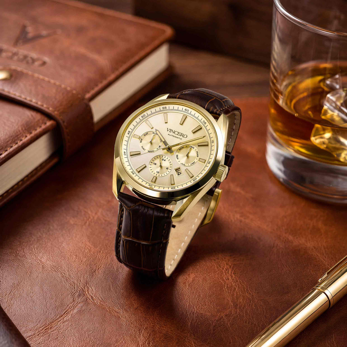 A Vincero wristwatch with a gold-tone case, cream dial, and dark brown leather strap, styled on a brown leather surface next to a glass of whiskey and a brown notebook.