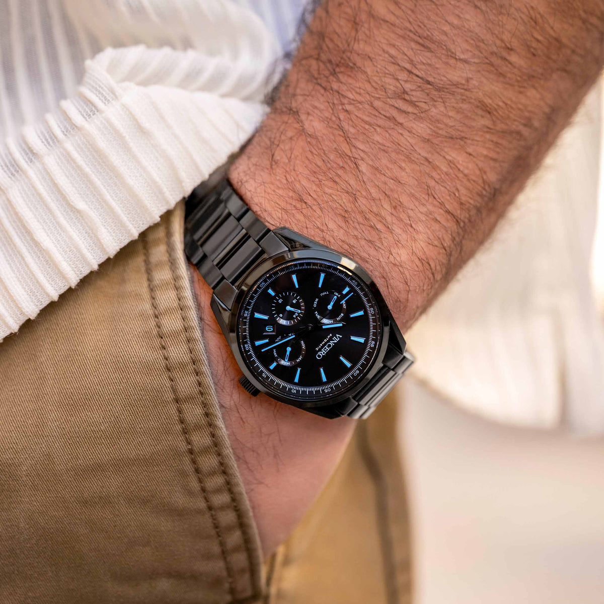 Black wristwatch with blue accents on a person's wrist, wearing a white shirt and beige pants.