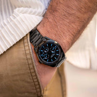 Black wristwatch with blue accents on a person's wrist, wearing a white shirt and beige pants.