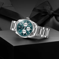 A stainless steel wristwatch with a teal blue dial and silver-tone metal bracelet, displayed diagonally on a dark gray block with black ribbon bows in the background.