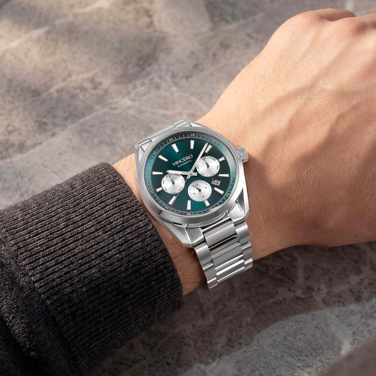 A wristwatch with a silver metal bracelet and a dark green dial, shown worn on a person's wrist against a textured gray background.
