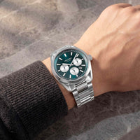 A wristwatch with a silver metal bracelet and a dark green dial, shown worn on a person's wrist against a textured gray background.
