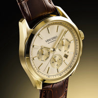 A Vincero Automatic watch with a polished gold-tone case, cream-colored dial with three subdials, and a dark brown leather strap, shown against a dark gradient background.