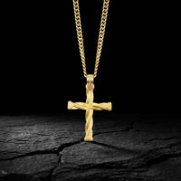 A gold cross pendant with a twisted, rope-like design hanging from a thick gold chain, displayed on a dark textured surface against a black background.