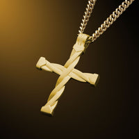 A gold cross pendant with a twisted, braided design detail, hanging from a gold curb chain against a dark brown gradient background.
