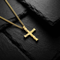 A gold cross pendant necklace with a twisted, draped design, resting on a textured black slate background.