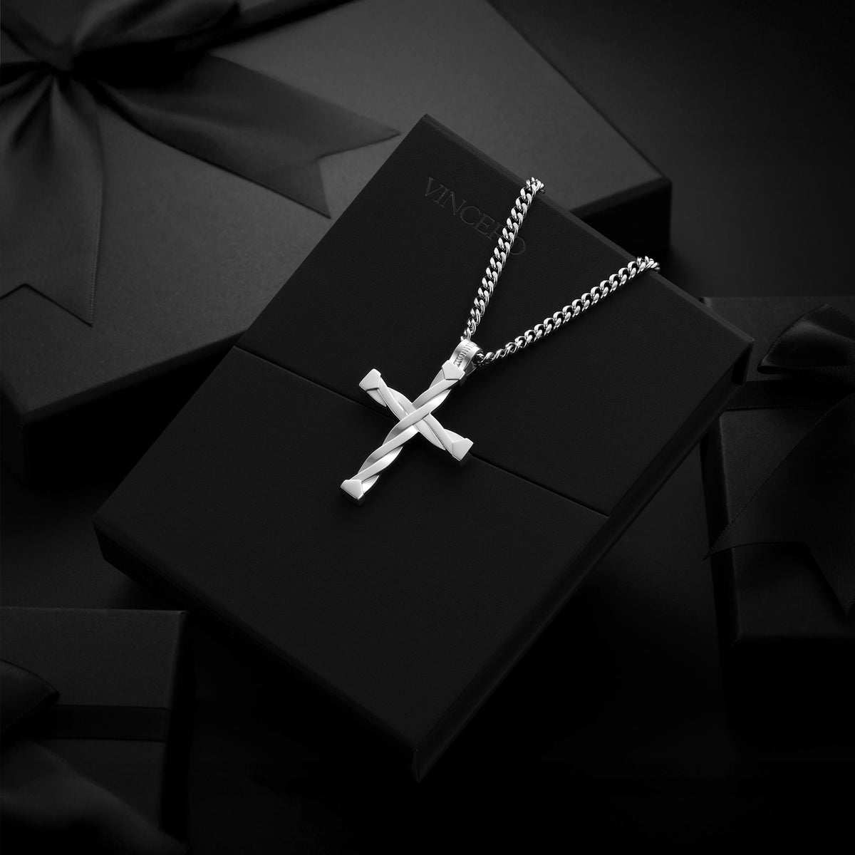 A silver cross pendant necklace on a silver chain, displayed on a black gift box with the brand name VINCI partially visible, set against a dark background with other black gift boxes and ribbons.