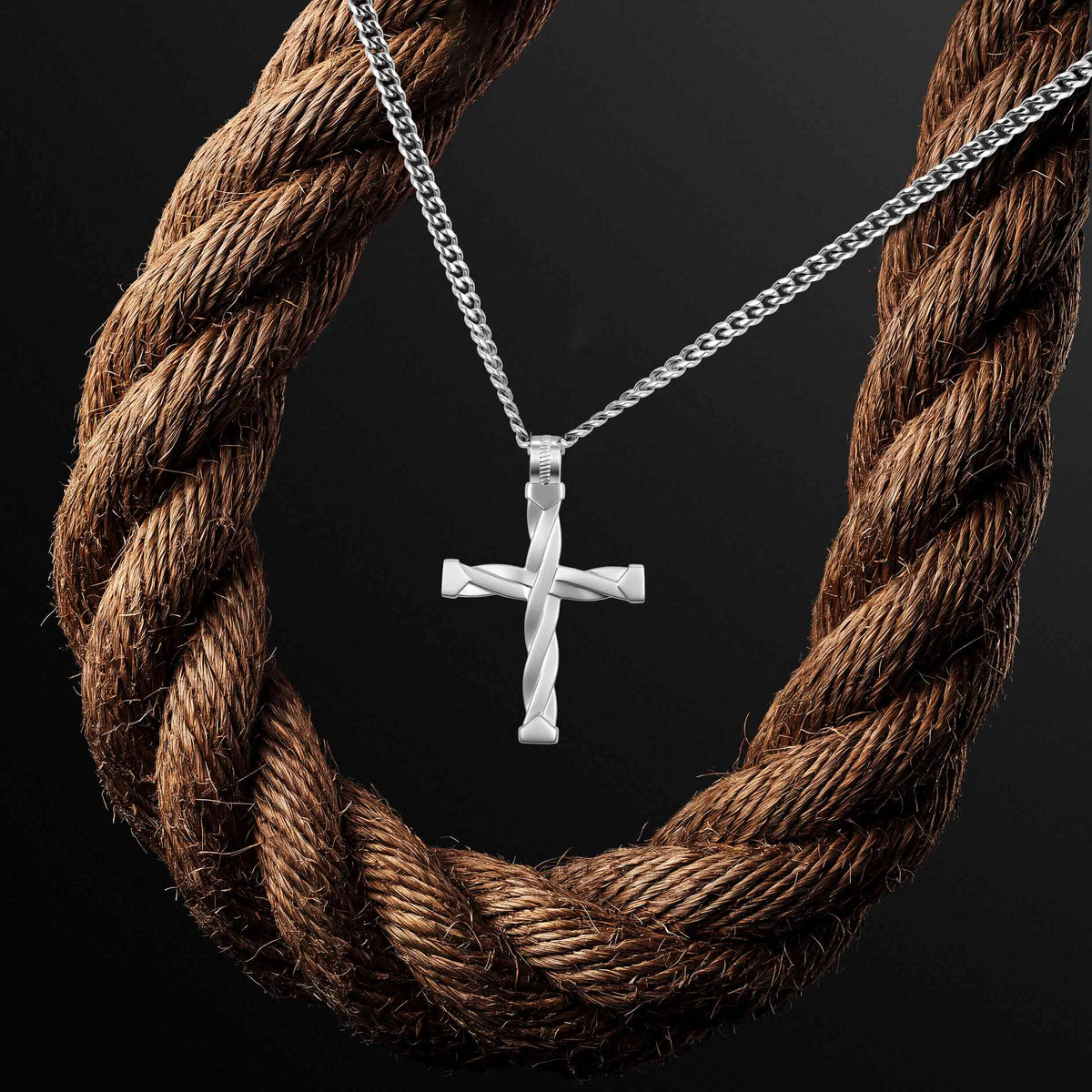 A silver cross pendant necklace with a twisted design, hanging from a silver chain, displayed against a dark background with a large brown rope prop.