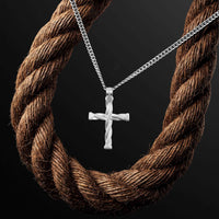 A silver cross pendant necklace with a twisted design, hanging from a silver chain, displayed against a dark background with a large brown rope prop.