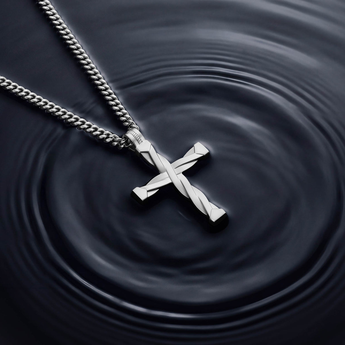 A silver cross pendant necklace with a twisted design, hanging from a silver curb chain against a dark background with concentric ripples.