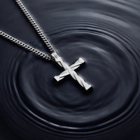 A silver cross pendant necklace with a twisted design, hanging from a silver curb chain against a dark background with concentric ripples.