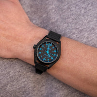 Black wristwatch with blue dial on a person's wrist against a gray background