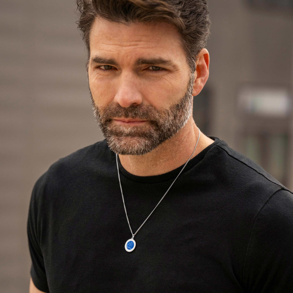 Man wearing a black t-shirt and necklace with a blue pendant against a blurred background