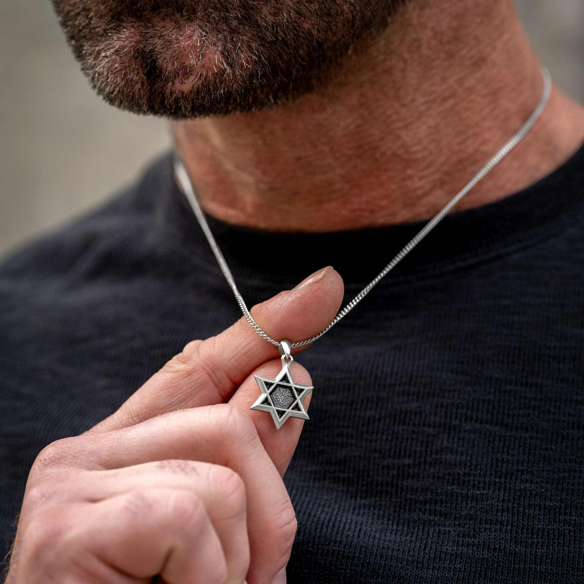 Man wearing silver chain necklace with silver Star of David pendant, using his finger to display the necklace
