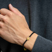 Hand wearing a black beaded bracelet with a gold cross charm against a dark background