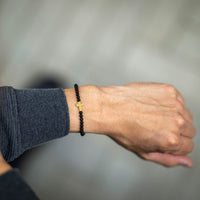 Person wearing a black beaded bracelet with a gold cross on a neutral background