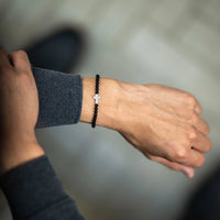 Person wearing a black beaded bracelet with a silver cross on a blurred background