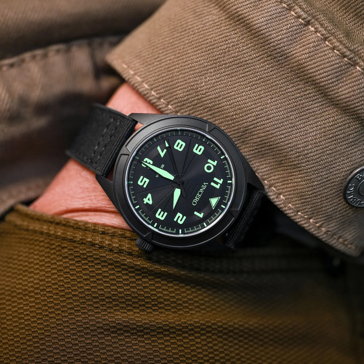 Black watch with green glowing face on a person's wrist against a brown fabric background