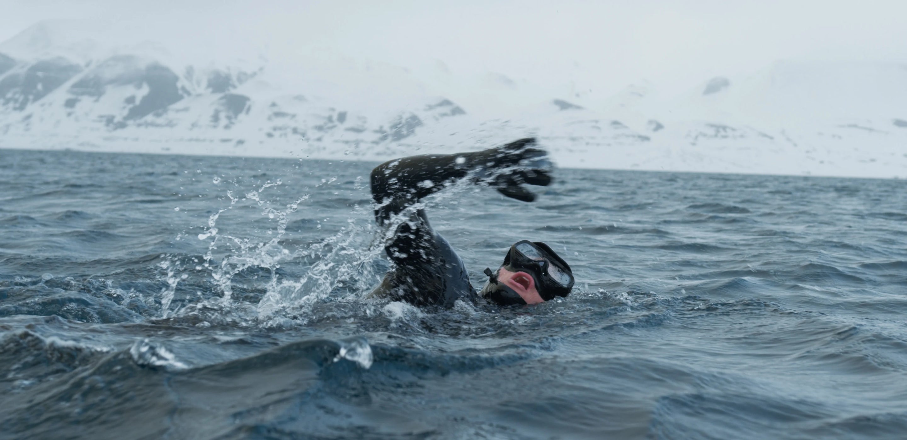 Man wearing a wetsuit swimming in the ocean with snow-covered mountains in the background.