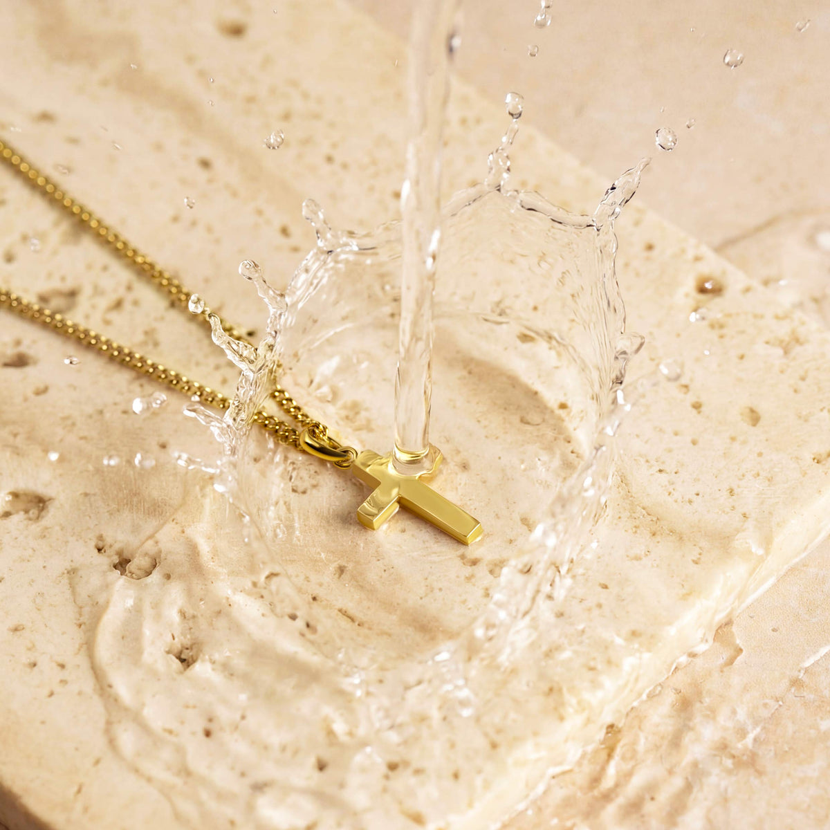 Gold necklace with a cross pendant on a textured beige surface with water droplets.