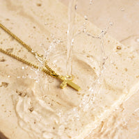 Gold necklace with a cross pendant on a textured beige surface with water droplets.