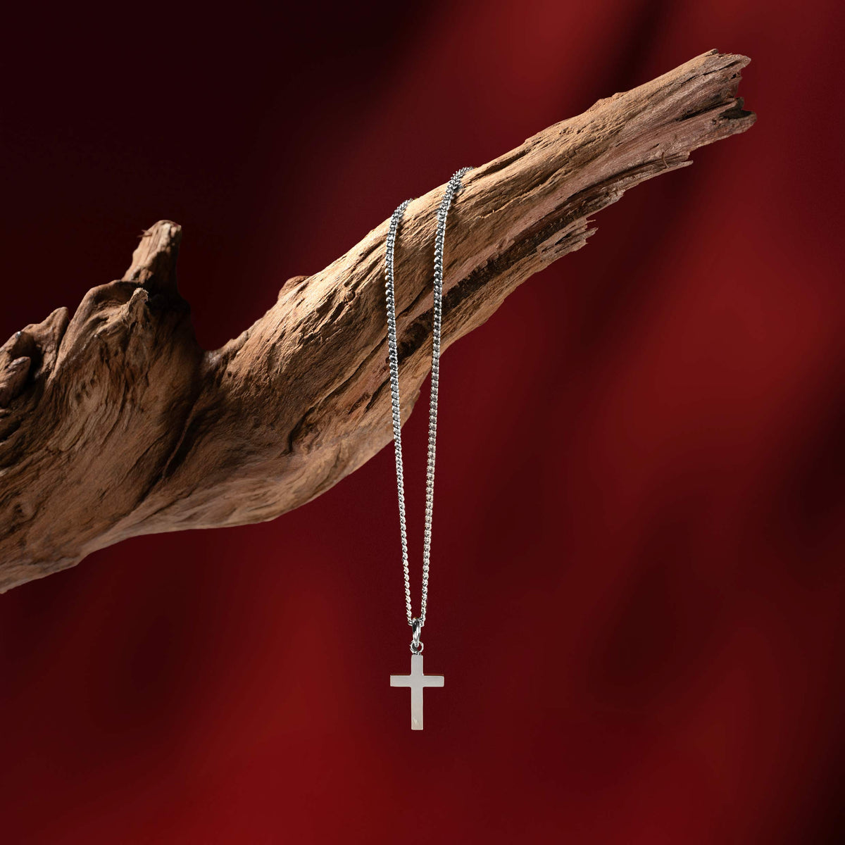 Silver necklace with a cross pendant on a wooden branch against a red background