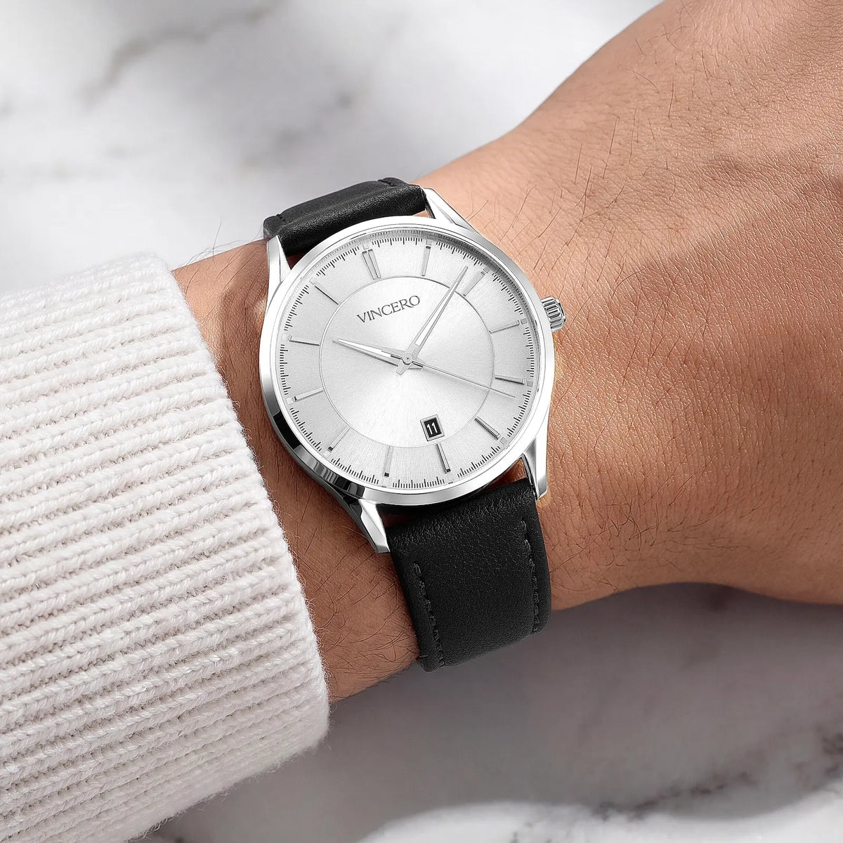 A Vincero wristwatch with a silver dial and a black leather strap, shown worn on a person's wrist with a white ribbed sweater sleeve visible.